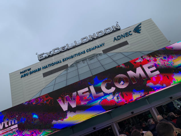 The exterior of the ExCeL London exhibition center, with a colorful digital welcome sign and the Abu Dhabi National Exhibitions Company branding. A crowd of attendees is gathered below the vibrant display.