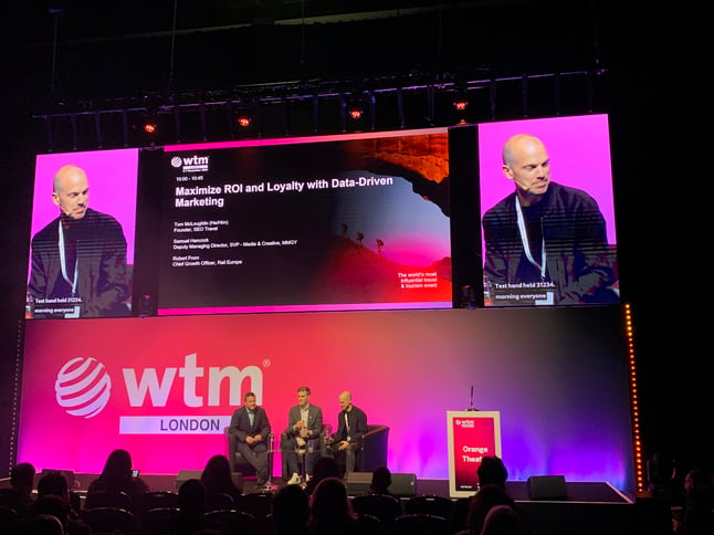 A panel discussion at WTM London 2024 on maximizing ROI and loyalty with data-driven marketing. Three speakers are seated on stage, with a large screen behind them displaying the panel details and a prominent WTM London logo