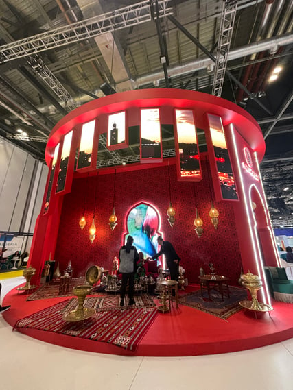 A striking red exhibition booth with golden lanterns and patterned rugs, featuring an archway with a digital screen displaying vivid visuals. People are interacting inside the booth, which is surrounded by decorative Middle Eastern-style furnishings at an indoor event.